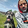 dog, puppy, person, man, hoodie, outdoor, grass, sky, clouds, stone_wall, beard, casual, collar, tongue_out, portrait, smiling, nature, friendship, leisure, daylight