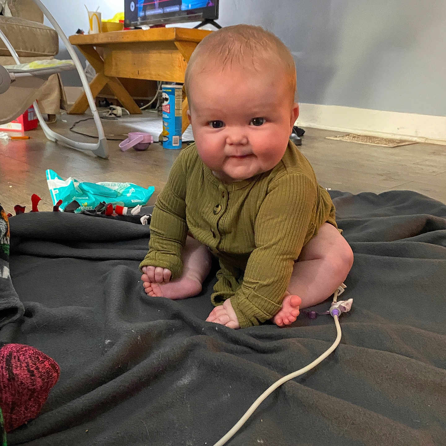 Rayleigh is registered to the contest to win money with this photo: baby, blanket, cable, casual, child, cozy, curious, face, floor, flooring, green_clothing, household_items, indoor, infant, room, sitting, smiling, television, toys, wooden_table