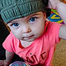 child, toddler, blue_eyes, green_hat, pink_shirt, smile_text, curious, indoor, person, portrait, baby, cute, expression, face, hand, clothing, playful, floor, colorful_background, closeup