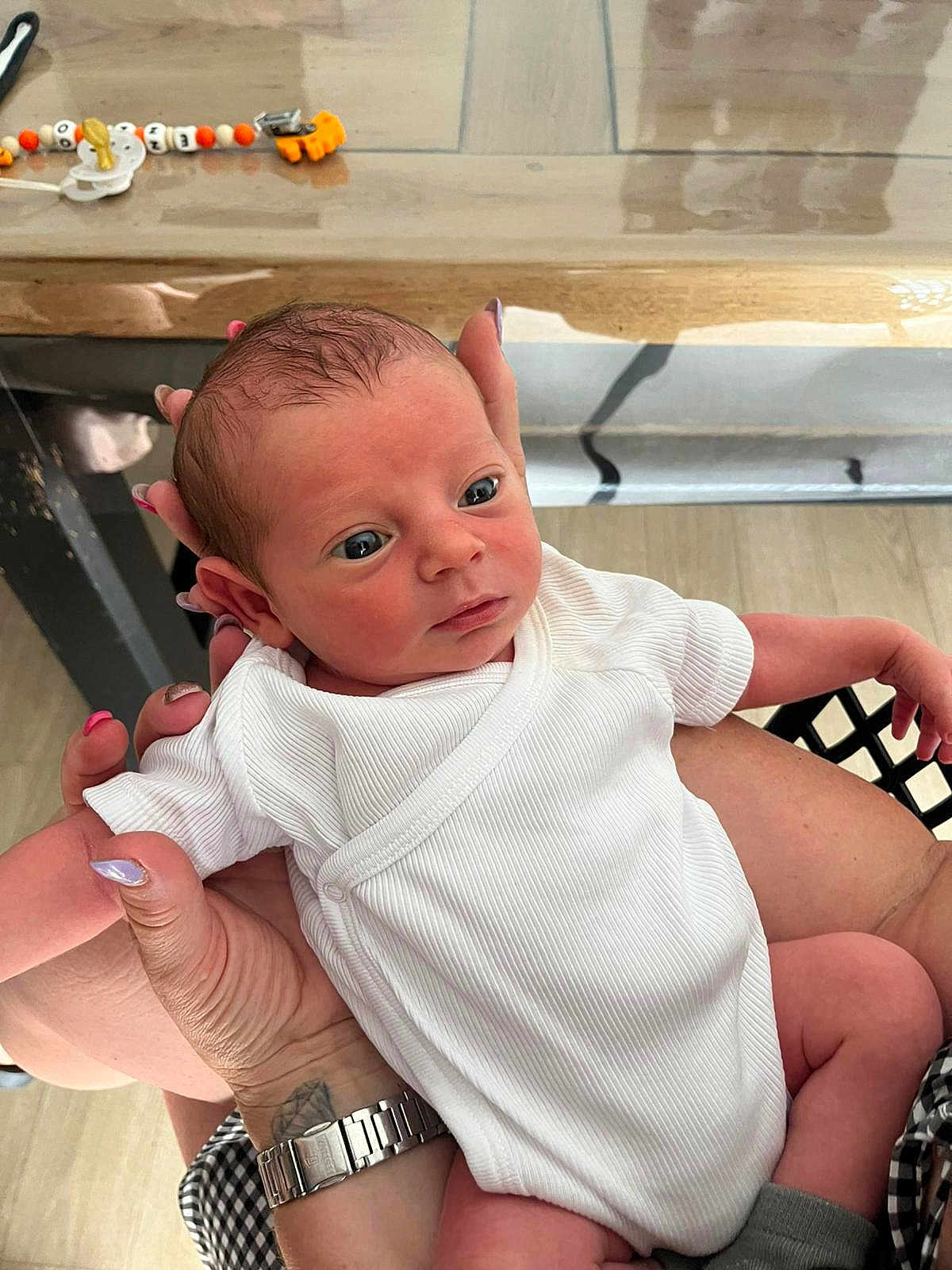 Ezzio a rejoint le concours — aidez-le/la à gagner de superbes lots ! newborn, baby, infant, white_onesie, hands, watch, nail_polish, pacifier_clip, wooden_table, indoor, person, skin, face, eyes, holding, curious, baby_clothing, closeup, human, arm