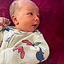 baby, infant, onesie, dinosaur_print, lying_down, red_blanket, soft_texture, curious_expression, face, head, hand, child, sleepwear, closeup, portrait, cute, newborn, skin, person, indoor