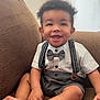 Josiah is registered to the contest to win money with this photo: child, toddler, smiling, bow_tie, suspenders, shorts, shirt, barefoot, couch, furniture, indoors, happy, cute, portrait, skin, hair, eyes, hands, feet, sitting