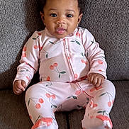 Adoree is registered to the contest to win money with this photo: baby, child, pink_onesie, cherry_pattern, sitting, couch, gray_couch, curly_hair, adorable, indoor, portrait, infant, clothing, cute, person, smiling, face, young_child, comfort, home