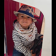 Julian participe au concours pour gagner de l'argent avec cette photo : child, hat, striped_clothing, scarf, steering_wheel, play_structure, indoor, smiling, jeans, sitting, person, casual_wear, cute, young, fun, playful, happy, portrait, hat_pattern, colorful