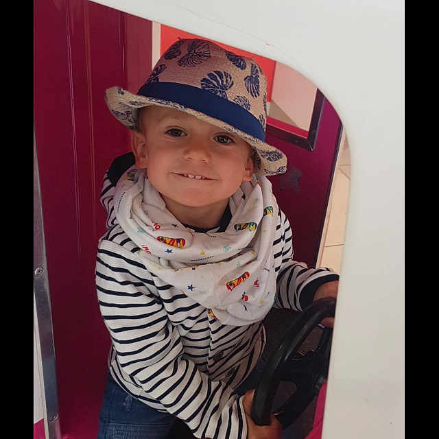 Julian participe au concours pour gagner de l'argent avec cette photo : casual_wear, child, colorful, cute, fun, happy, hat, hat_pattern, indoor, jeans, person, play_structure, playful, portrait, scarf, sitting, smiling, steering_wheel, striped_clothing, young