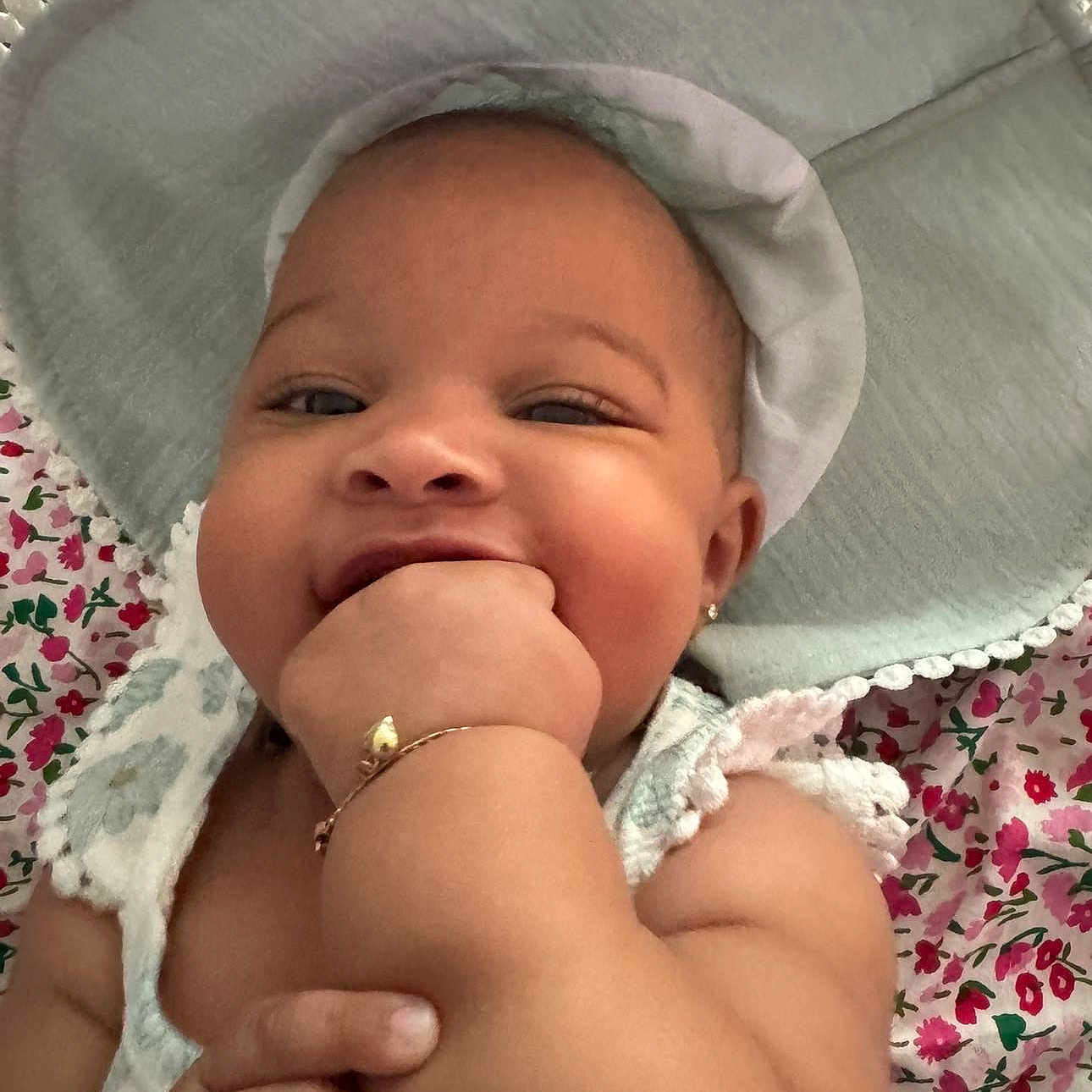 Kayomi is registered to the contest to win money with this photo: accessory, adorable, arm, baby, bracelet, chewing, child, chubby, closeup, cute, cute_expression, floral_blanket, hand, happy, hat, infant, lying_down, portrait, skin, smiling