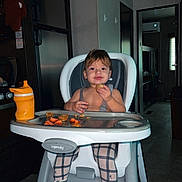 Jaxon is registered to the contest to win money with this photo: toddler, child, high_chair, snack, kitchen, food, sippy_cup, legs, feet, plaid_pants, indoor, person, face, happy, eating, tray, baby, footrest, hair, room