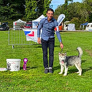 Alpha a rejoint le concours — aidez-le/la à gagner de superbes lots ! man, dog, husky, grass, outdoor, park, event, leash, blue_shirt, black_pants, sunny, tent, banner, car, greenery, pet, animal, smile, competition, rosette