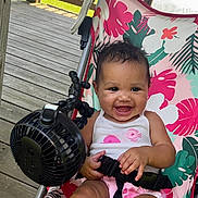 Arielle joined the competition — help win amazing prizes! baby, child, smiling, stroller, seatbelt, fan, wooden_deck, outdoor, lake, floral_pattern, pink_clothing, white_tank_top, happy, cute, summer, sunlight, person, infant, seated, playful
