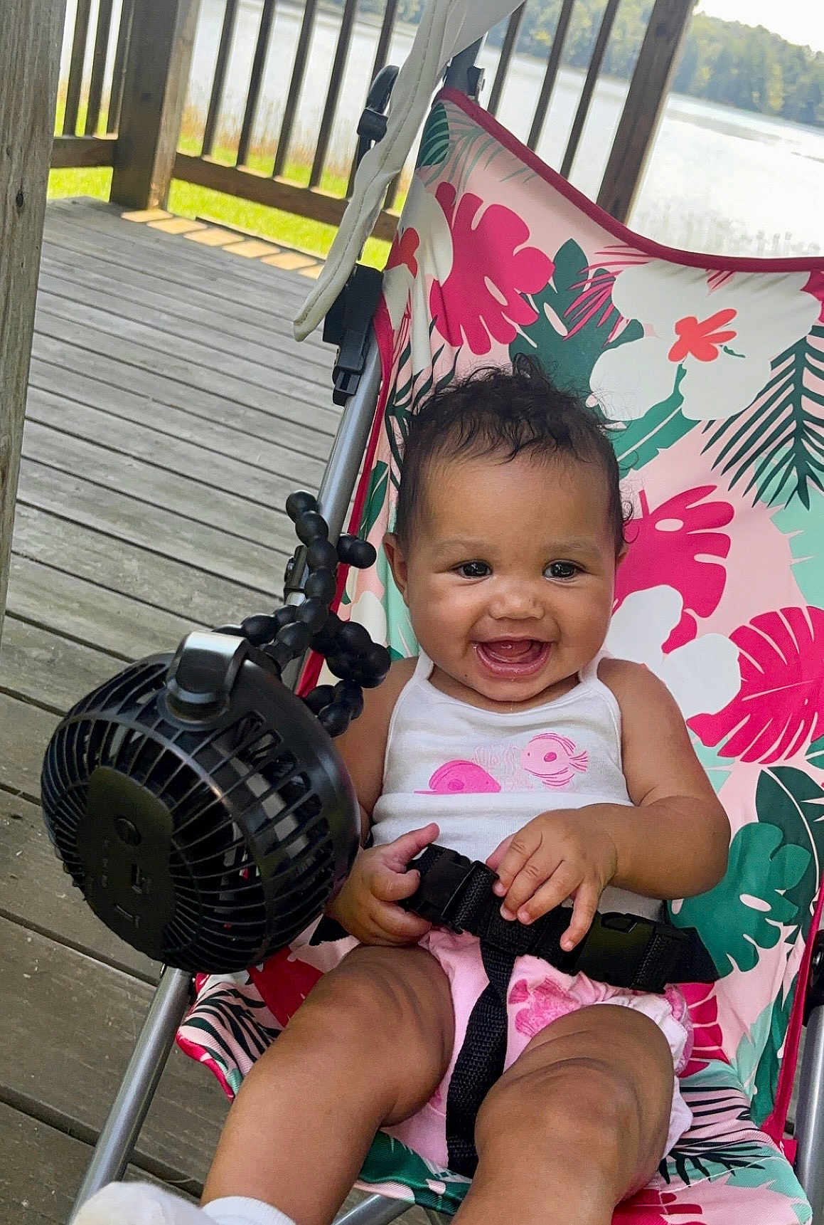 Arielle joined the competition — help win amazing prizes! baby, child, smiling, stroller, seatbelt, fan, wooden_deck, outdoor, lake, floral_pattern, pink_clothing, white_tank_top, happy, cute, summer, sunlight, person, infant, seated, playful
