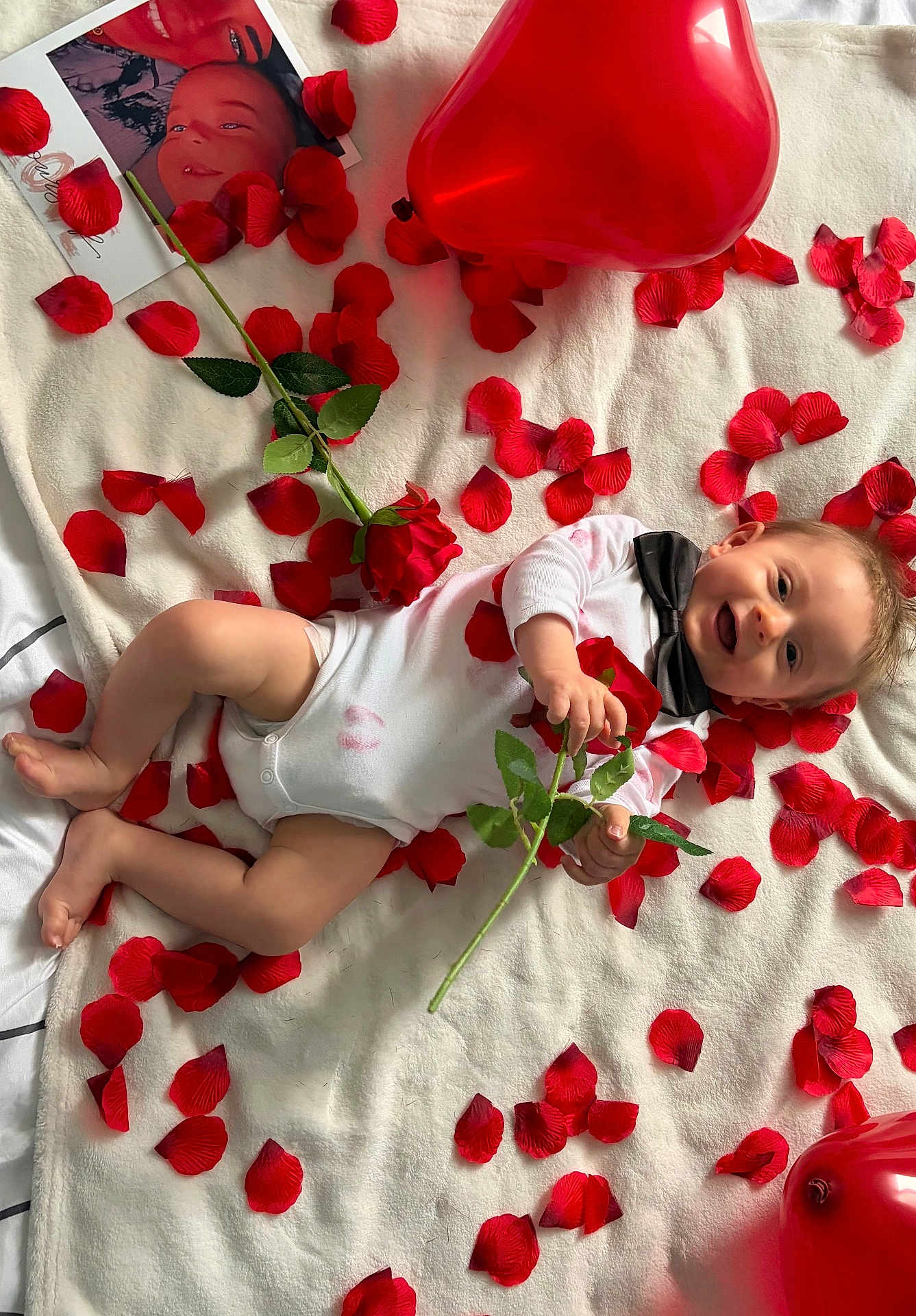 Isaak participe au concours pour gagner de l'argent avec cette photo : baby, infant, smiling, rose, rose_petals, balloon, heart_balloon, blanket, bow_tie, onesie, lipstick_mark, printed_photo, portrait, lying_down, playful, red_color, white_blanket, hands, feet, scattered_petals