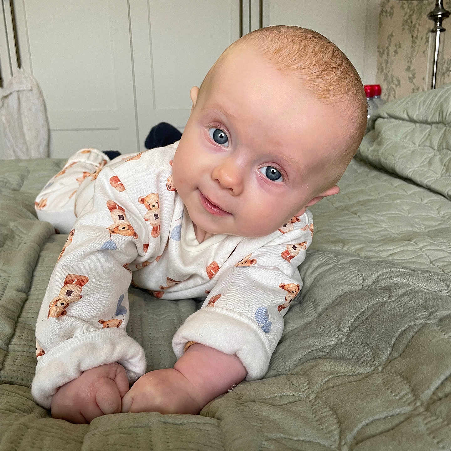 Tommy is registered to the contest to win money with this photo: baby, bed, bedroom, blue_eyes, child, cozy, crib, cute, gaze, hands, head_tilt, indoor, infant, neutral_background, portrait, quilt, sleepwear, smile, soft_lighting, teddy_bear_onesie