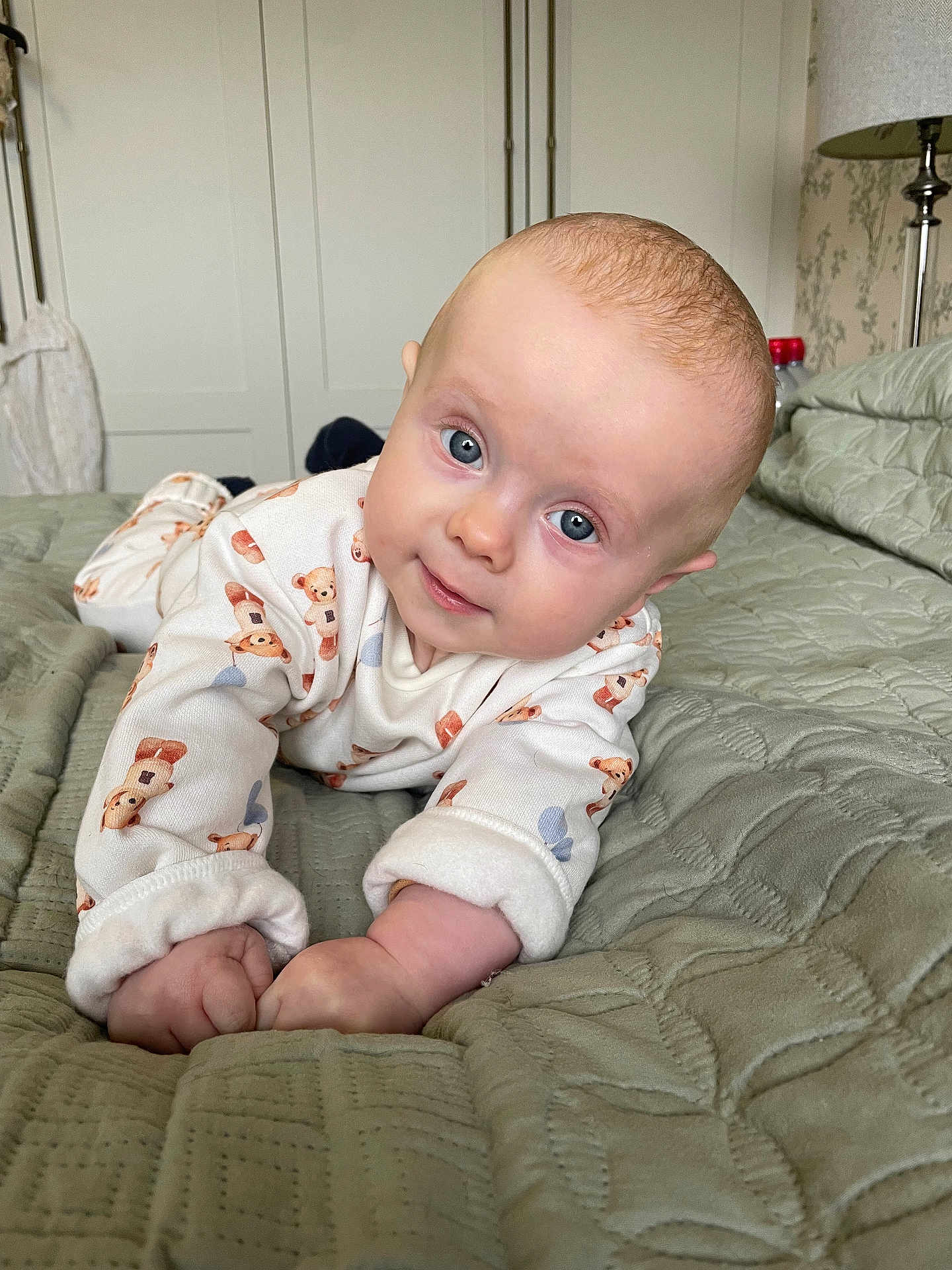 Tommy is registered to the contest to win money with this photo: baby, infant, blue_eyes, teddy_bear_onesie, bed, quilt, indoor, child, cute, portrait, head_tilt, hands, cozy, soft_lighting, gaze, neutral_background, sleepwear, bedroom, crib, smile