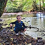 child, boy, river, rocks, water, trees, forest, nature, outdoor, crouching, smiling, clothing, shoes, daylight, greenery, play, exploration, riverbank, small_object, leaves