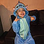 child, blue_onesie, costume, stitch, smiling, couch, indoor, playful, happy, hand_gesture, hooded, facial_expression, young, person, casual, cozy, fun, portrait, home, furniture