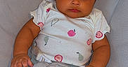 Majesty is registered to the contest to win money with this photo: baby, child, infant, portrait, sitting, cushioned_seat, white_background, curly_hair, brown_skin, looking_at_camera, onesie, baby_clothing, fruit_pattern, pink_details, blue_pants, hands, legs, indoors, close_up, adorable