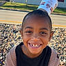 child, smiling, portrait, outdoor, sunny, paper_cup, balancing, playful, happy, face, teeth, black_shirt, cardigan, clothing, pebbles, street, building, window, grass, photography