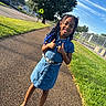 child, smiling, outdoor, sidewalk, street, denim_dress, braids, hairstyle, thumbs_up, portrait, full_body, sunny, summer, neighborhood, grass, tree, crosswalk_sign, fence, houses, fashion