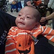 Natalia is registered to the contest to win money with this photo: baby, blurred_background, casual_wear, child, clothing, cute, face, hand, indoor, infant, orange_sweater, people, person_holding, portrait, pumpkin_design, shopping_mall, smile, striped_clothing, tattoo, toddler