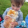 child, kid, sandwich, food, picnic, blanket, grass, park, outdoor, eating, plastic_wrap, hoodie, casual_clothing, cute, small_child, young, nature, daytime, funny, happy