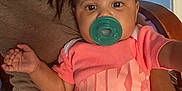Caliyah is registered to the contest to win money with this photo: baby, pacifier, pink_clothing, person, child, indoor, sitting, head, face, arm, hair, furniture, blue_wall, wooden_chair, adult, holding, casual_wear, curly_hair, closeup, expression