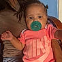 baby, pacifier, pink_clothing, person, child, indoor, sitting, head, face, arm, hair, furniture, blue_wall, wooden_chair, adult, holding, casual_wear, curly_hair, closeup, expression