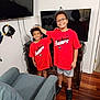 child, smiling, indoor, red_shirt, glasses, television, headphones, wood_floor, furniture, chair, happy, person, sibling, shorts, black_shoes, technology, cable, console, playful, curly_hair