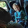 toddler, child, car_seat, smile, happy, seatbelt, car_interior, window, sunlight, clothing, person, safety, car, headrest, seat, curly_hair, green_shirt, blue_pants, daylight, vehicle