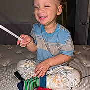 Isaiah is registered to the contest to win money with this photo: toddler, child, smiling, sitting, mattress, blue_shirt, striped_shirt, colorful_socks, laughing, indoor, playful, hand, legs_crossed, curly_hair, young_child, happy, person, bedroom, toy, cable