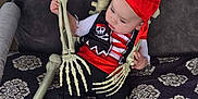 Jeremiah joined the competition — help win amazing prizes! baby, infant, child, skeleton, pirate_costume, red_bandana, hat, bones, ribcage, skull_prop, chair, cushion, patterned_blanket, bare_feet, toes, hands, playful, holiday_decoration, costume_party, portrait