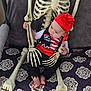 baby, infant, child, skeleton, pirate_costume, red_bandana, hat, bones, ribcage, skull_prop, chair, cushion, patterned_blanket, bare_feet, toes, hands, playful, holiday_decoration, costume_party, portrait
