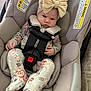 baby, bow, car_seat, child, cute, fabric, floral_clothing, footed_pajamas, gray, headband, indoor, infant, padded_seat, person, seatbelt, serious_expression, sitting, small_hands, straps, toddler