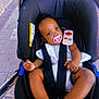 Noa-Rai joined the competition — help win amazing prizes! baby, stroller, pacifier, toy_remote, child, infant, seatbelt, outdoor, sidewalk, pavement, black_stroller, white_clothing, cute, small_hands, bare_feet, curly_hair, infant_seat, relaxed, daylight, portrait