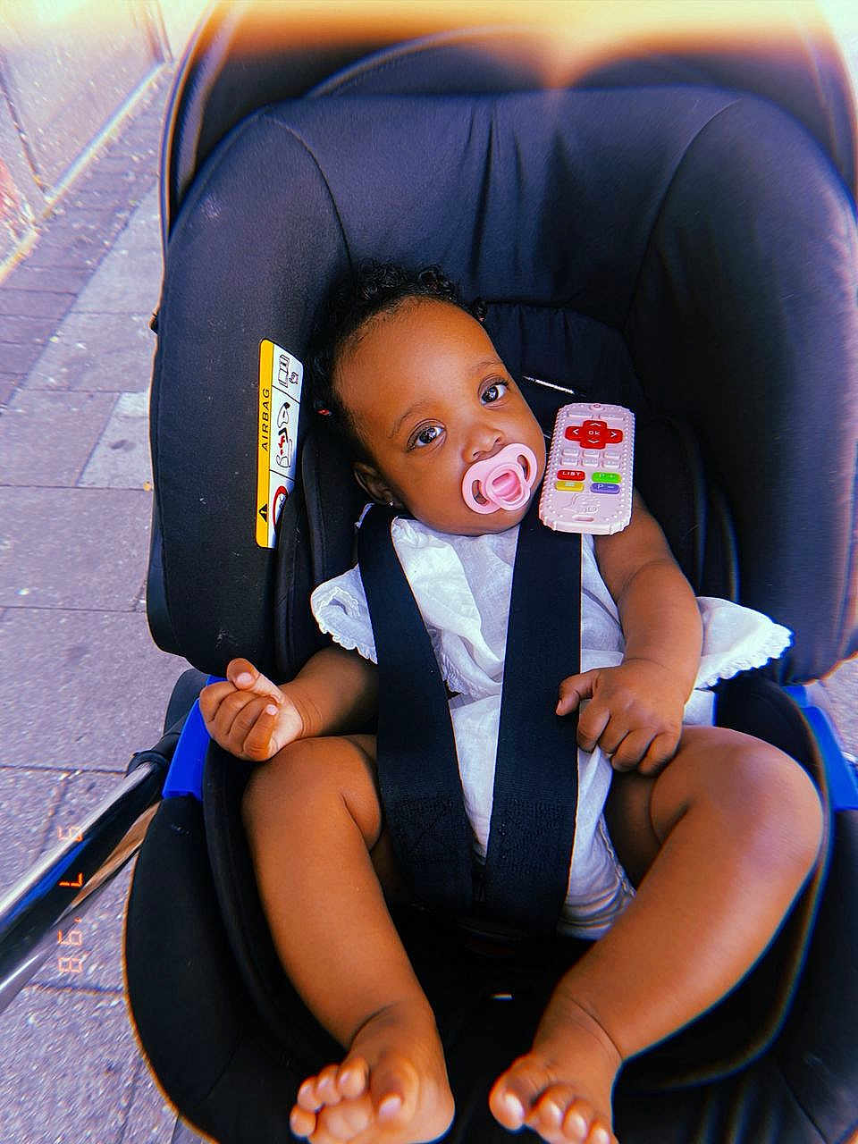 Noa-Rai joined the competition — help win amazing prizes! baby, stroller, pacifier, toy_remote, child, infant, seatbelt, outdoor, sidewalk, pavement, black_stroller, white_clothing, cute, small_hands, bare_feet, curly_hair, infant_seat, relaxed, daylight, portrait