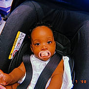 Noa-Rai is registered to the contest to win money with this photo: baby, pacifier, car_seat, child, black_hair, earrings, white_clothing, infant, seatbelt, indoor, store, floor, hand, curious, face, person, closeup, safety, child_seat, cute