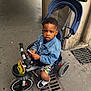 child, clothing, concrete, denim_jacket, expression, grate, handlebar, lollipop, outdoor, person, shoes, shorts, sidewalk, street, toddler, toy, tricycle, urban, vehicle, wheel