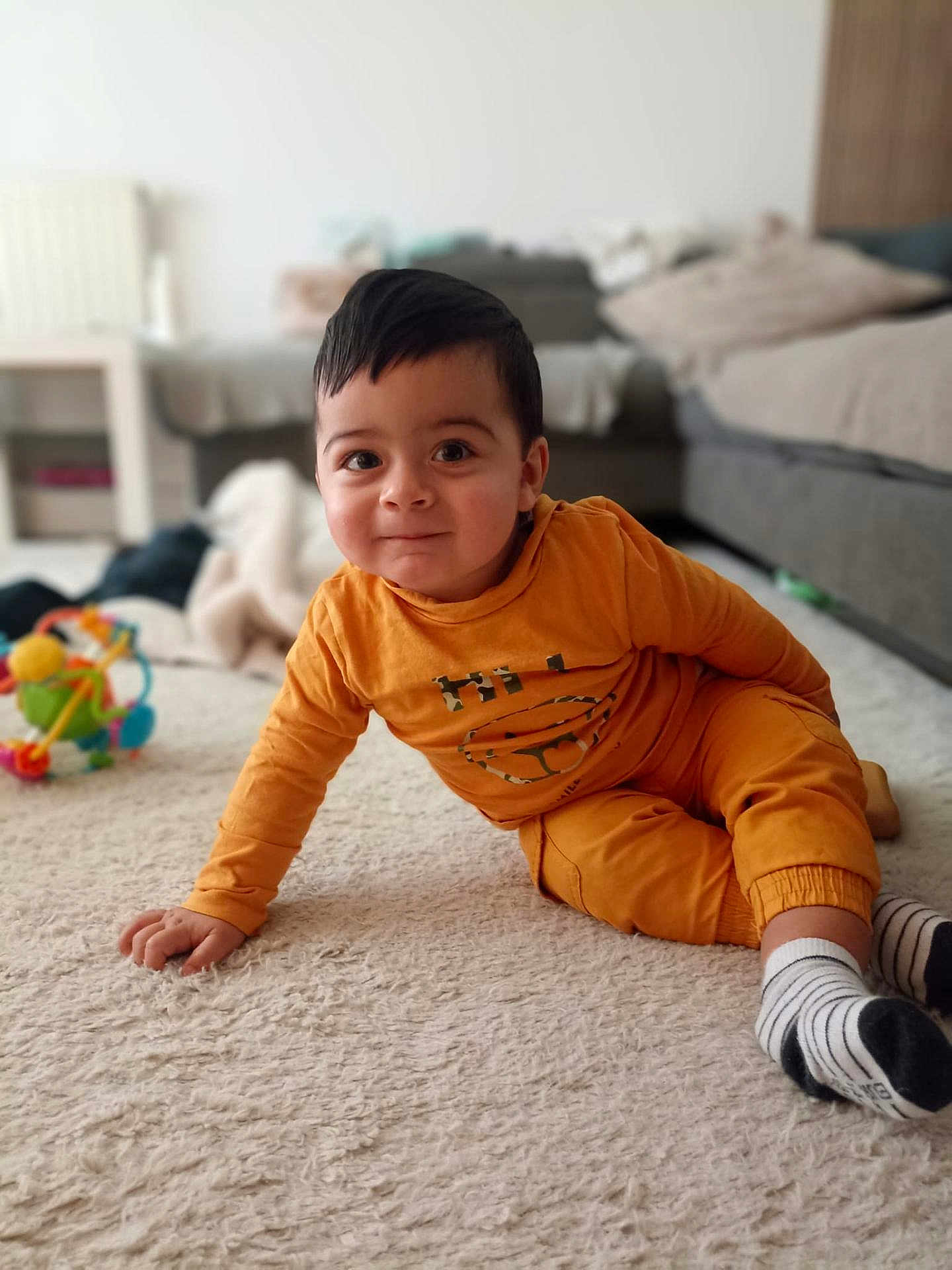 Jayro a rejoint le concours — aidez-le/la à gagner de superbes lots ! carpet, child, couch, curious, cute, face, floor, furniture, hair, home, indoor, orange_clothing, person, playful, relaxed, smile, socks, toddler, toy, young_child