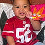 Kavika joined the competition — help win amazing prizes! toddler, child, red_jersey, nfl_logo, number_52, blue_jeans, quilted_blanket, indoor, seated, curious_expression, hand_in_mouth, short_hair, brown_skin, fabric_texture, colorful_background, casual_clothing, portrait, face, person, cute