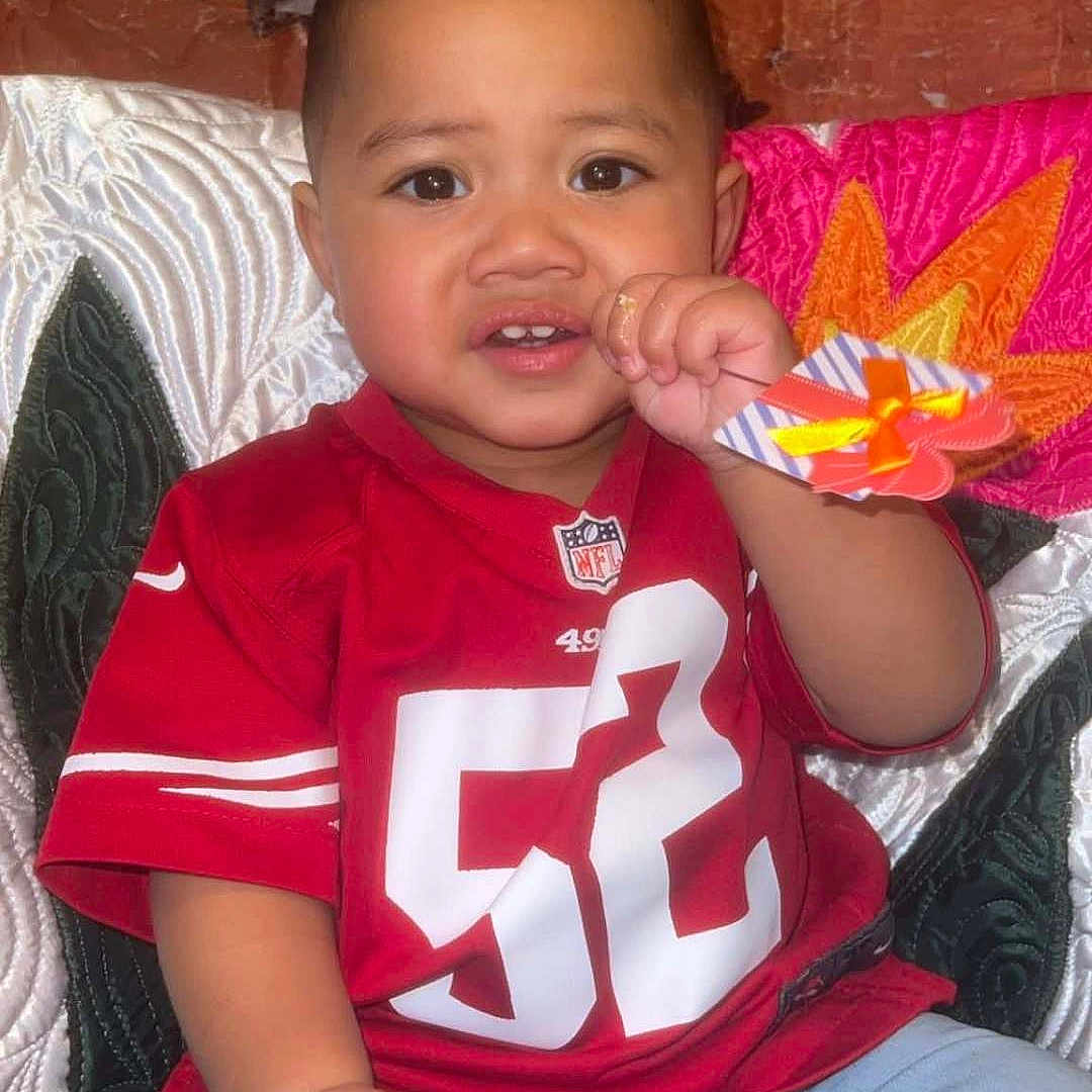 Kavika joined the competition — help win amazing prizes! blue_jeans, brown_skin, casual_clothing, child, colorful_background, curious_expression, cute, fabric_texture, face, hand_in_mouth, indoor, nfl_logo, number_52, person, portrait, quilted_blanket, red_jersey, seated, short_hair, toddler