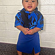 Kavika joined the competition — help win amazing prizes! toddler, child, boy, blue_shirt, skirt, sandals, standing, indoor, carpet, brick_wall, smiling, hands_clasped, short_hair, cute, portrait, person, fashion, happy, casual, young_child