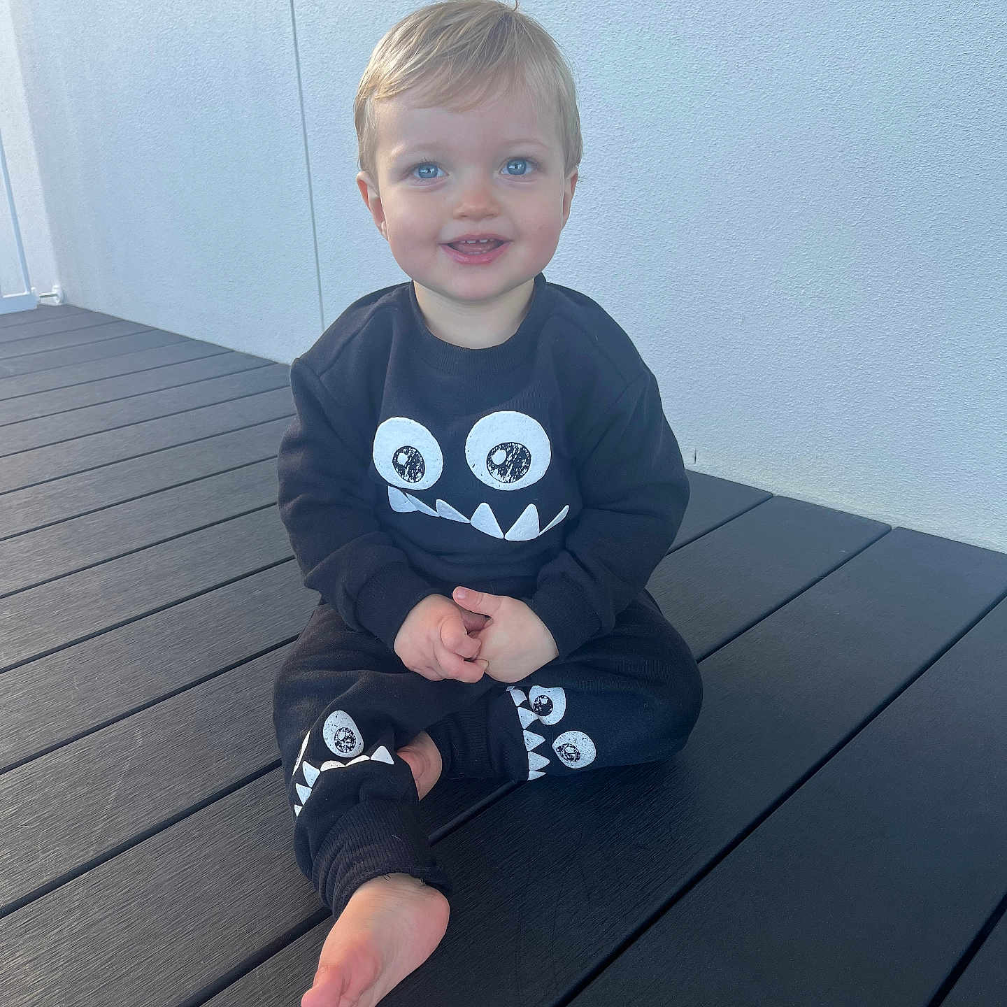Kyson is registered to the contest to win money with this photo: barefoot, black_clothing, blue_eyes, casual, child, cute, daylight, happy, long_sleeves, monster_face, outdoor, pants, person, playful_design, sitting, smiling, toddler, wall, wooden_deck, young_child