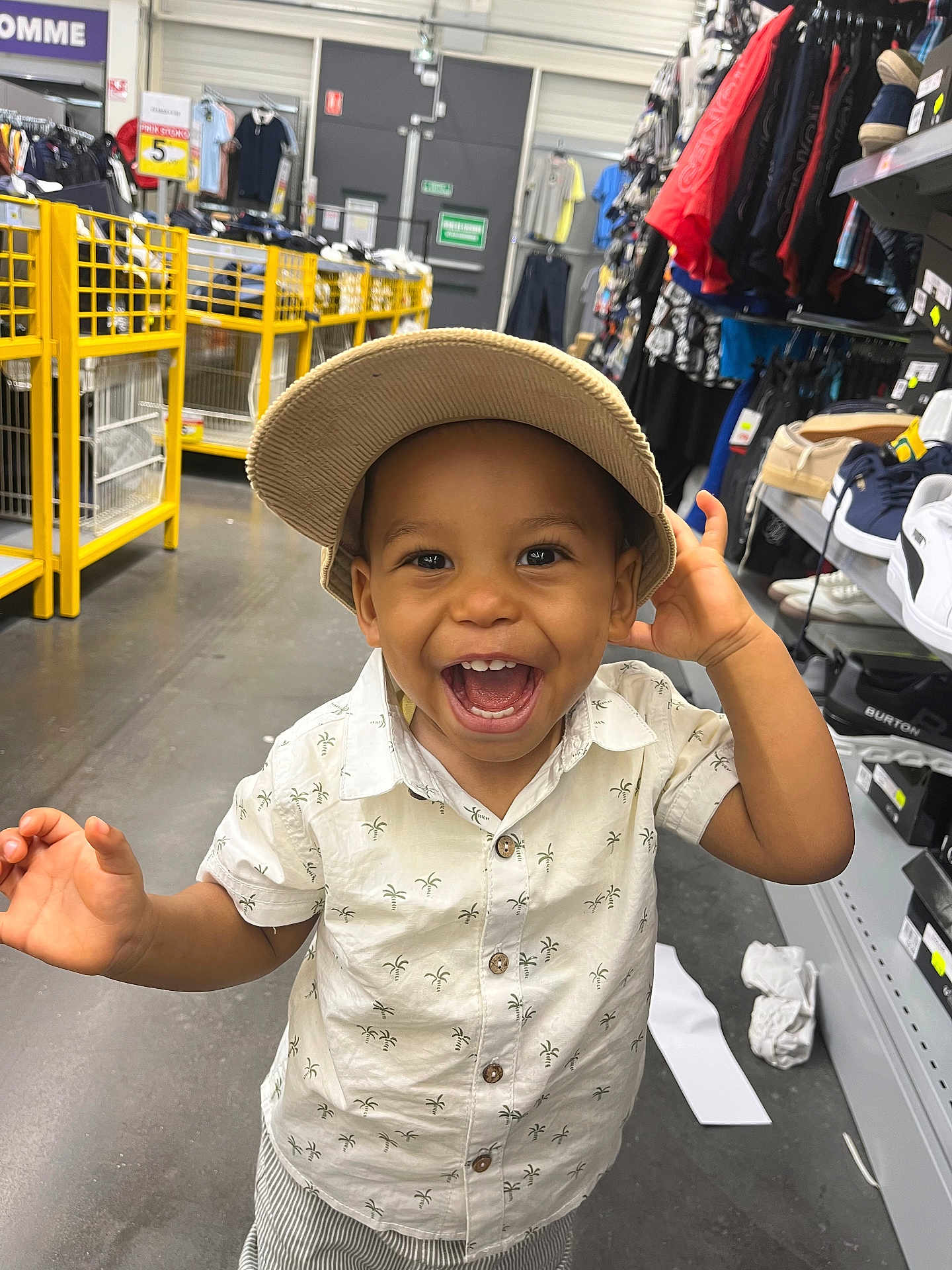 Samuel participe au concours pour gagner de l'argent avec cette photo : toddler, child, smiling, hat, beige_cap, white_shirt, patterned_shirt, shopping_store, clothing_rack, shoes, happy, portrait, indoor, floor, casual_clothing, boy, joyful, cute, retail, aisle