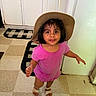 child, toddler, girl, cowboy_hat, pink_shirt, smiling, portrait, indoor, kitchen, floor, tile_floor, cabinet, door, rug, sandals, standing, short_hair, dark_hair, looking_at_camera, playful