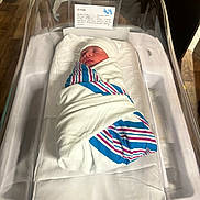 Emylain is registered to the contest to win money with this photo: newborn, baby, hospital, bassinet, blanket, swaddled, sleeping, infant, hat, bed, medical, indoors, person, rest, peaceful, white, blue, striped, soft, quiet