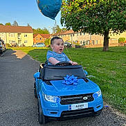 Azari is registered to the contest to win money with this photo: toddler, child, toy_car, blue_car, balloon, birthday, outdoor, grass, tree, sidewalk, residential_area, sunny_day, happy, celebration, blue_shirt, small_vehicle, license_plate, front_view, excited, playtime