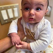 Silas is registered to the contest to win money with this photo: baby, infant, child, person, hand, holding, white_clothing, teddy_bear_print, face, eyes, cute, indoor, light_switch, skin, expression, portrait, sitting, soft_light, closeup, curious