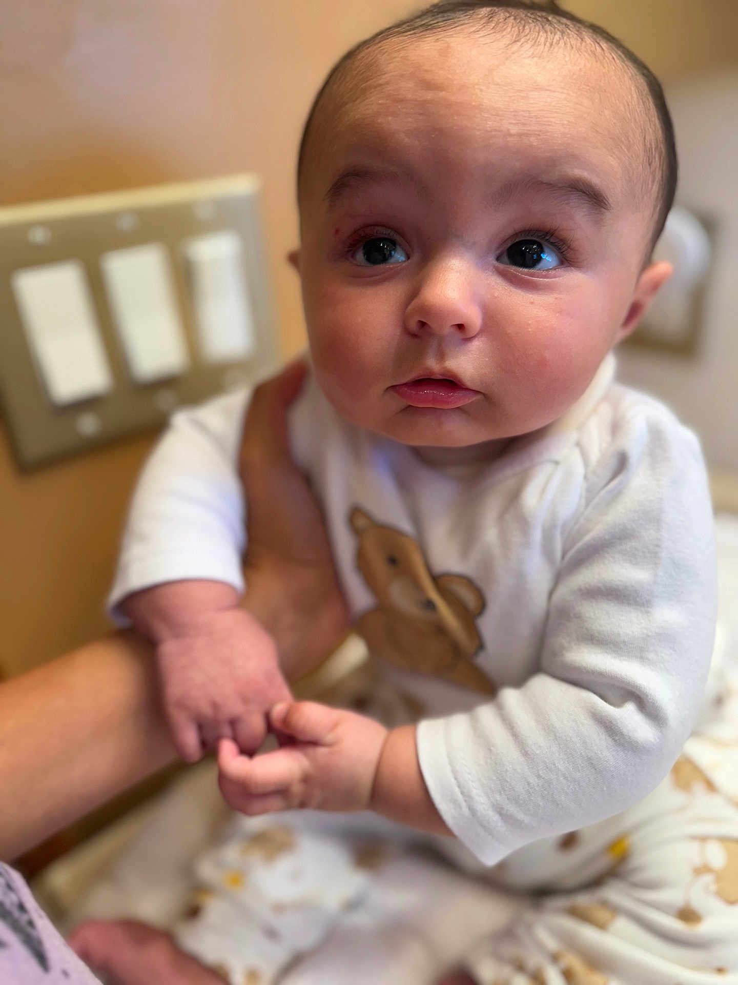 Silas is registered to the contest to win money with this photo: baby, infant, child, person, hand, holding, white_clothing, teddy_bear_print, face, eyes, cute, indoor, light_switch, skin, expression, portrait, sitting, soft_light, closeup, curious