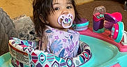 Eliana is registered to the contest to win money with this photo: toddler, pacifier, baby_walker, child, indoor, toy, playpen, floor, carpet, clothing, hair, face, eyes, furniture, room, childcare, colorful, seat, infant, curious