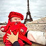 baby, infant, child, red_coat, red_beret, smiling, happy, eiffel_tower, paris, landmark, graffiti, stone_ledge, hands, adult_hand, outdoor, cloudy_sky, portrait, travel, tourist, hat