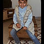 blue_shirt, boy, carpet, casual_clothing, child, curly_hair, furniture, happy, home, indoor, jeans, person, portrait, puffer_vest, rug, sitting, smiling, sneakers, television, wooden_bench